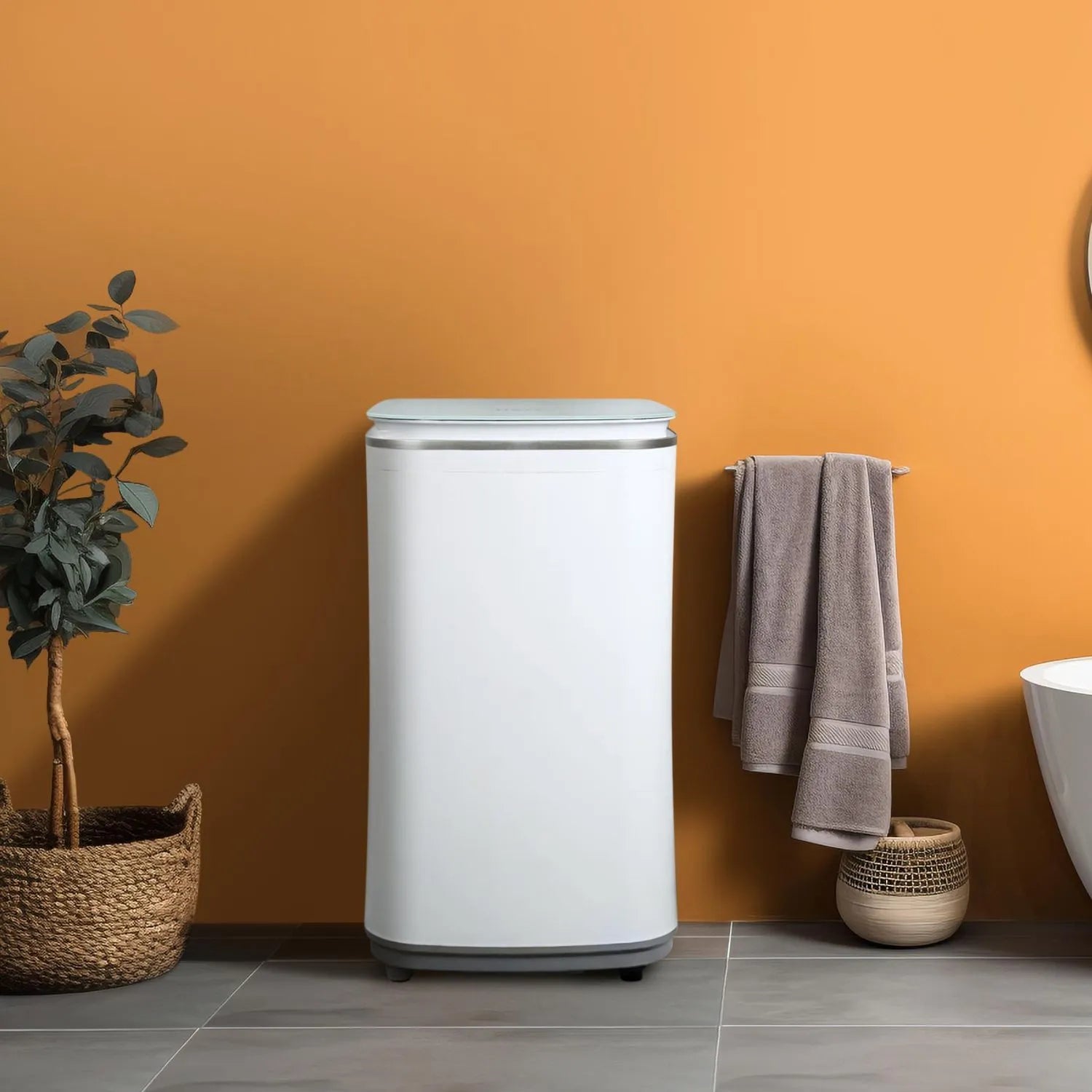 HAVA tvättmaskin från Go-clean.se placerad mot varm orange vägg i badrum, omgiven av frodig växt i rottingkruka, hängande handduk och fristående badkar som skapar en inbjudande atmosfär.